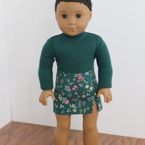 May include: A doll wearing a green long-sleeved shirt and a green and floral print skirt. The doll has brown skin and black hair in a bun.