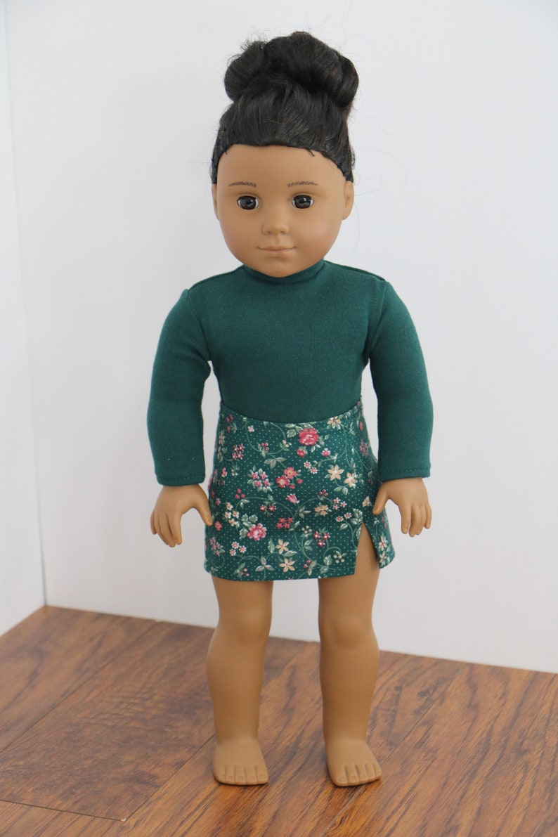 May include: A doll wearing a green long-sleeved shirt and a green and floral print skirt. The doll has brown skin and black hair in a bun.
