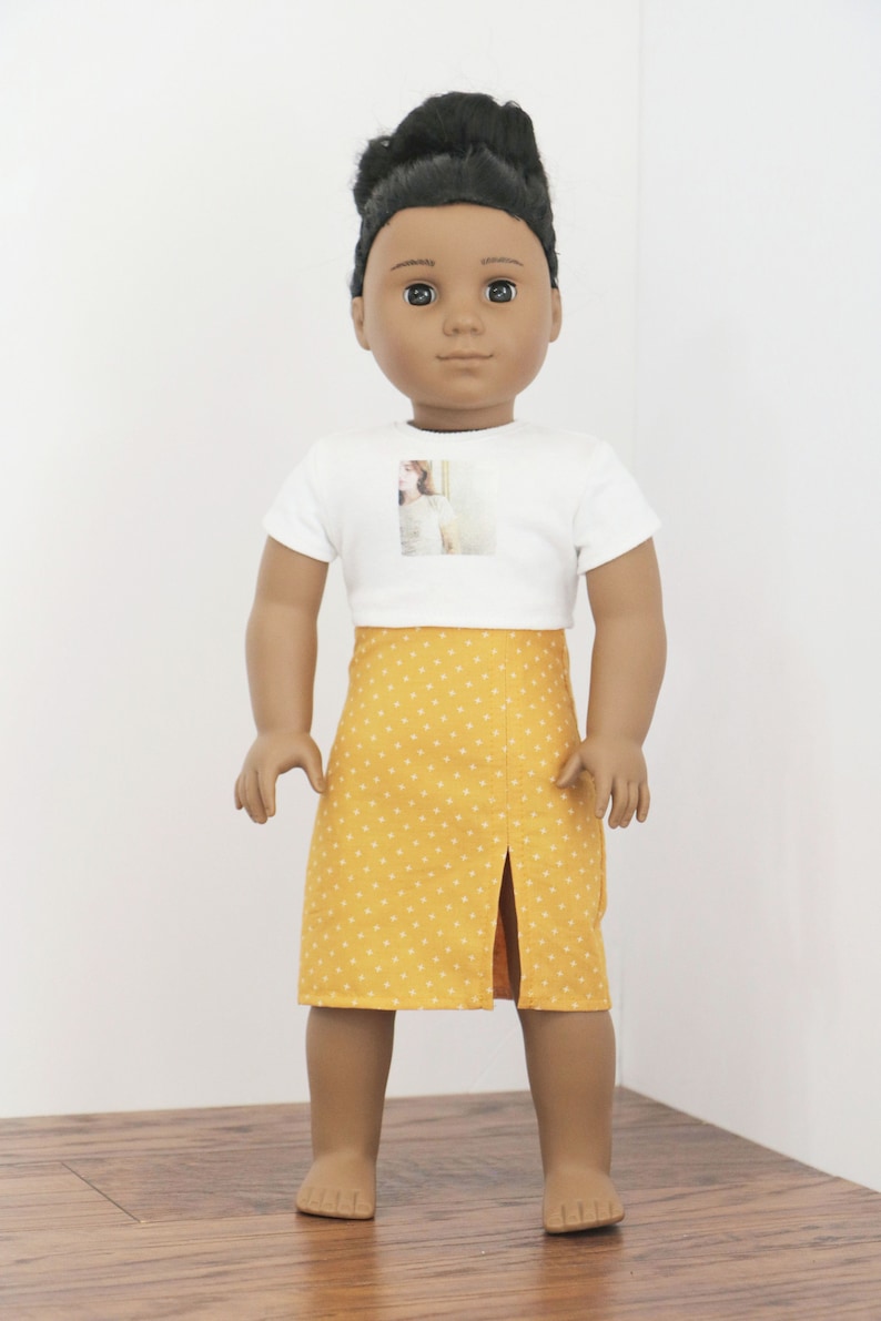 May include: A doll wearing a white t-shirt with a photo printed on the front and a yellow skirt with white polka dots.