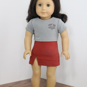 May include: A doll wearing a grey short-sleeved shirt with the text "Ron Jon Surf Shop" and red shorts. The doll is wearing red sunglasses and has brown hair.