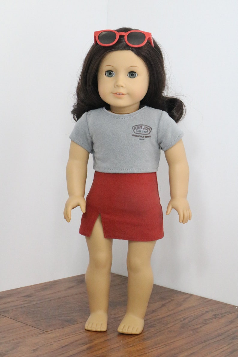 May include: A doll wearing a grey short-sleeved shirt with the text "Ron Jon Surf Shop" and red shorts. The doll is wearing red sunglasses and has brown hair.