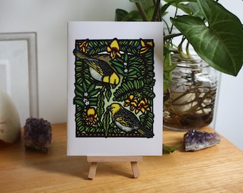 May include: A greeting card with a vibrant illustration of two birds surrounded by green leaves and yellow flowers. The artwork has a black border and is displayed on a small wooden easel. The background includes plants and decorative items, creating a natural aesthetic.