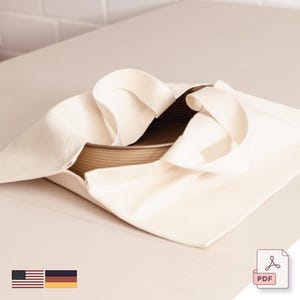 Puede incluir: Una bolsa de lona beige con un recipiente marrón en su interior. La bolsa tiene un diseño sencillo con una base rectangular y un asa. El recipiente parece ser una bandeja redonda para hornear. La imagen está tomada desde un ángulo ligeramente elevado.