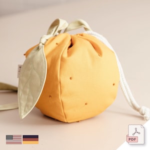 Puede incluir: Una bolsa de cordón de tela amarilla con forma de naranja con un detalle de hoja verde. La bolsa tiene un cordón blanco y está hecha de una tela suave y texturizada.
