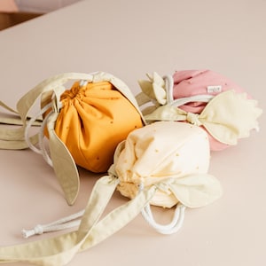 May include: Three fabric drawstring pouches shaped like fruit, in mustard yellow, pale yellow, and dusty pink. Each pouch features small embroidered dots, light green leaf-shaped accents, and a white braided rope closure for securing the opening.