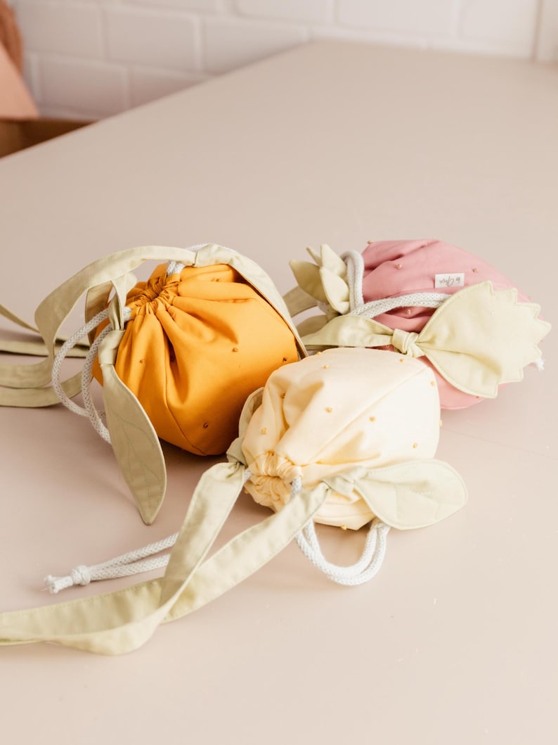 May include: Three fabric drawstring pouches shaped like fruit, in mustard yellow, pale yellow, and dusty pink. Each pouch features small embroidered dots, light green leaf-shaped accents, and a white braided rope closure for securing the opening.