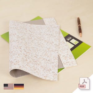 May include: A white and beige fabric folder with a floral pattern. The folder is folded in half and has a grey fabric lining. The folder is sitting on a wooden surface.
