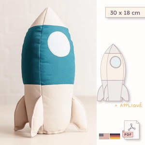 May include: A plush rocket ship toy featuring a cream-colored nose and base with a teal midsection and a circular white window. The rocket measures 30 x 18 cm. A digital diagram shows the rocket outline with an appliqué and a PDF file icon.