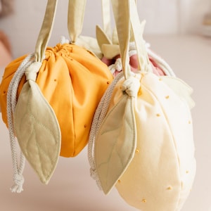 May include: Two fabric drawstring pouches shaped like fruit, one bright orange and one pale yellow. Each pouch features light green fabric leaves attached to the top, a white braided drawstring closure, and small decorative beads on the surface.