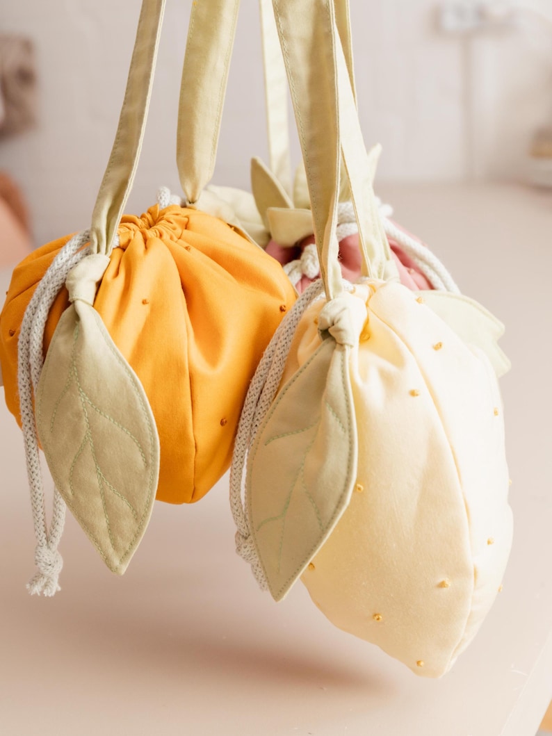 May include: Two fabric drawstring pouches shaped like fruit, one bright orange and one pale yellow. Each pouch features light green fabric leaves attached to the top, a white braided drawstring closure, and small decorative beads on the surface.