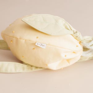May include: A pale yellow drawstring bag shaped like a lemon, featuring a quilted light green leaf accent on top, small yellow embroidered dots across the surface, white rope drawstrings, and a small white fabric tag labeled by Ofe&ccedil;a.
