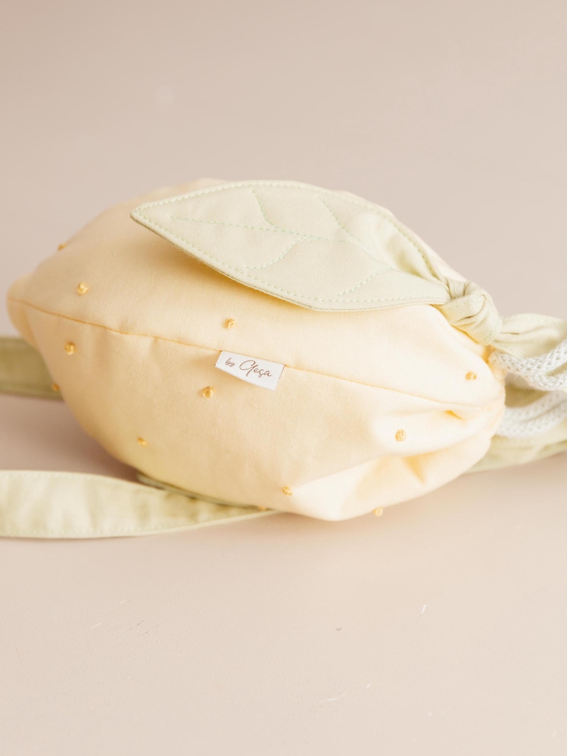 May include: A pale yellow drawstring bag shaped like a lemon, featuring a quilted light green leaf accent on top, small yellow embroidered dots across the surface, white rope drawstrings, and a small white fabric tag labeled by Ofe&ccedil;a.