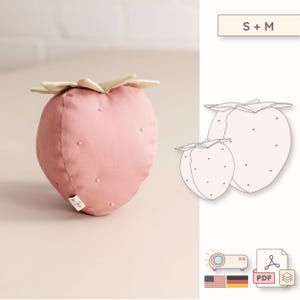 Puede incluir: Un juguete de peluche con forma de fresa en rosa empolvado con un tallo verde claro y pequeños puntos bordados. La imagen incluye un dibujo lineal de dos tamaños de fresa etiquetados como S + M, un icono de proyector y un icono de PDF con banderas.