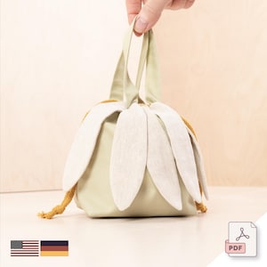 Puede incluir: Una bolsa con cordón verde claro con un diseño de flor de margarita. La bolsa tiene un cuerpo verde claro, pétalos color crema y un cordón mostaza. La bolsa está sujeta por una mano. Un accesorio único.
