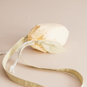 May include: A pale yellow drawstring pouch shaped like a fruit, featuring small yellow dots across the surface. The bag includes a light green fabric leaf accent near the top and a long, matching light green shoulder strap with white cord.