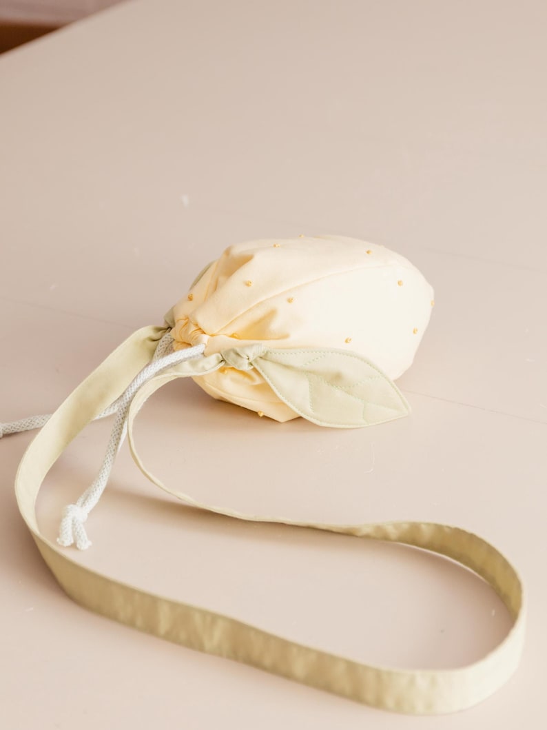 May include: A pale yellow drawstring pouch shaped like a fruit, featuring small yellow dots across the surface. The bag includes a light green fabric leaf accent near the top and a long, matching light green shoulder strap with white cord.