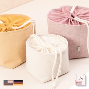 May include: Three fabric drawstring bags in yellow, cream, and pink. Each bag has a contrasting interior and a white drawstring closure. The bags are made of a textured fabric and are arranged on a light-coloured surface.