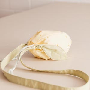 May include: A pale yellow drawstring pouch shaped like a lemon, featuring small yellow bead accents across the surface. The bag includes a light green fabric leaf detail attached to the top and a long, matching light green shoulder strap.