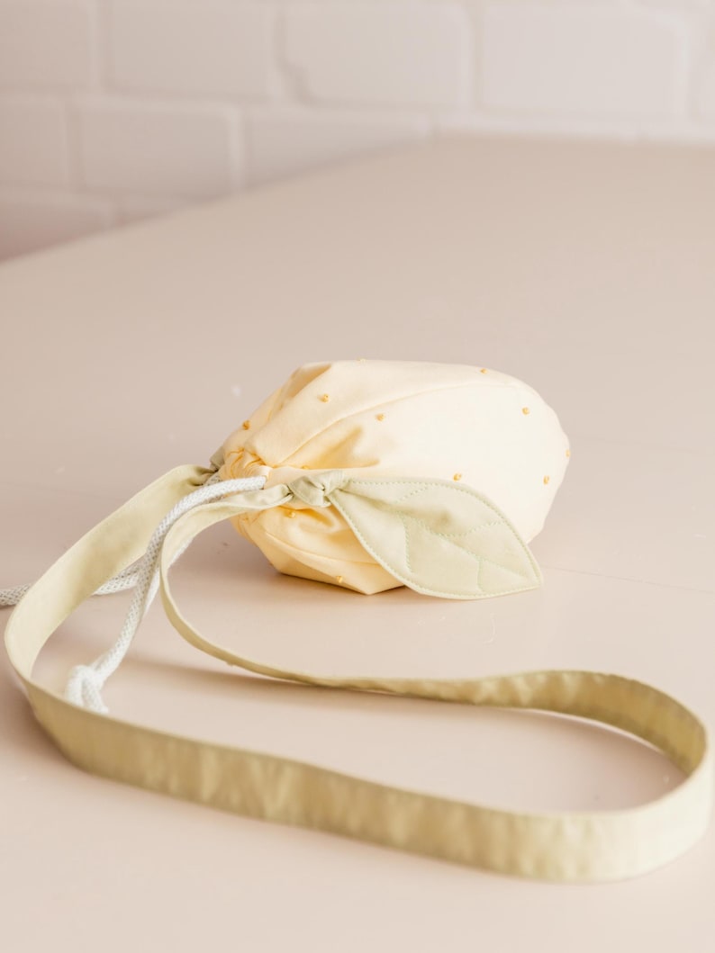 May include: A pale yellow drawstring pouch shaped like a lemon, featuring small yellow bead accents across the surface. The bag includes a light green fabric leaf detail attached to the top and a long, matching light green shoulder strap.