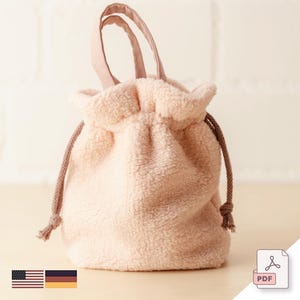 Op de afbeelding: Een zachte, roze, pluche trekkoordtas met een bruin koord en een roze stoffen handvat. De tas is gemaakt van een pluche materiaal en heeft een geplooide bovenkant. De tas staat op een wit oppervlak.