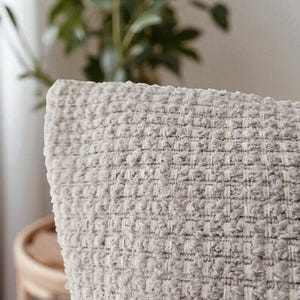 Peut inclure: Gros plan d'un coussin texturé gris clair. Le tissu présente un motif tissé en forme de gaufre. Le coussin fait partie d'une collection de décoration intérieure, suggérant confort et style pour les espaces de vie.