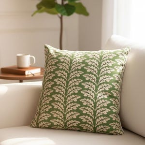 May include: A square scatter cushion featuring a repeating pattern of cream-coloured fern-like leaves on a solid green background. The fabric has a visible weave texture, and the cushion is positioned on a plain, light-coloured upholstered surface.