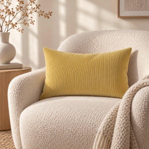May include: A rectangular throw cushion in a solid mustard yellow colour featuring a vertical ribbed texture. The cushion is positioned centrally on a cream-coloured textured armchair, showcasing a consistent fabric pattern across its entire surface.