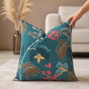 Könnte beinhalten: Ein quadratisches Dekokissen mit einem blaugrünen Hintergrund und einem botanischen Druck. Das Muster zeigt stilisierte Blätter in Braun- und Dunkelblautönen sowie Gruppen kleiner rosa Blüten und blauer Beeren auf dünnen weißen Zweigen.