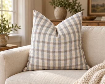 May include: A square throw pillow featuring a classic buffalo check pattern in muted shades of blue-gray and off-white. The fabric has a visible woven texture, and the pillow is plumply filled, creating a soft, structured shape on a sofa.