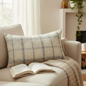 May include: A cozy living room scene featuring a beige sofa with a decorative pillow and a knitted throw. An open book rests on the sofa. A white mug sits on a small wooden side table. A fireplace is in the background.
