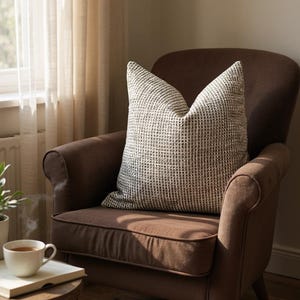 May include: A brown armchair with a patterned cushion in neutral tones. A small wooden side table holds a cup of tea and a book. The room is lit by natural light filtering through a sheer curtain.