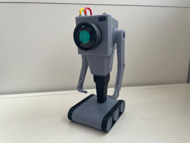 Puede incluir: Un juguete robot gris con una lente de c&aacute;mara como cara, brazos articulados y una base de orugas. El robot tiene tres cables de colores en la parte superior y est&aacute; hecho de pl&aacute;stico. El juguete mide aproximadamente 15 cm de altura.