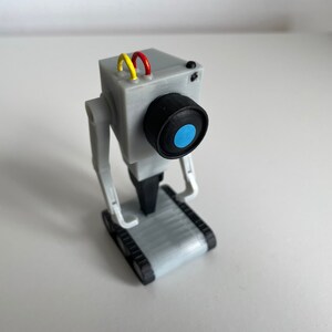 Puede incluir: Un juguete robot gris con una lente de c&aacute;mara y un centro azul. El robot tiene una cabeza rectangular con cables amarillos y rojos, y brazos y piernas articulados. Est&aacute; montado sobre una base con orugas.
