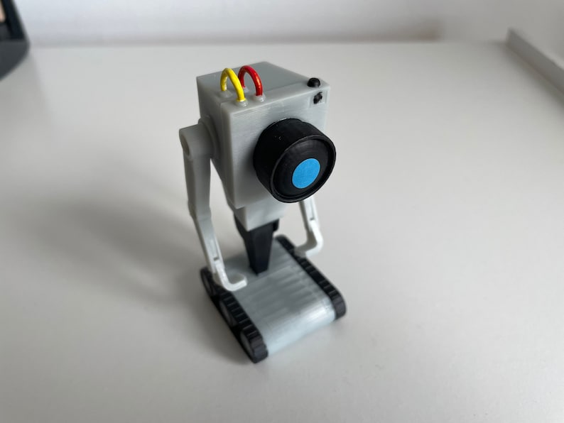 Puede incluir: Un juguete robot gris con una lente de c&aacute;mara y un centro azul. El robot tiene una cabeza rectangular con cables amarillos y rojos, y brazos y piernas articulados. Est&aacute; montado sobre una base con orugas.
