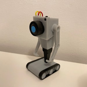 Puede incluir: Un juguete robot gris impreso en 3D con una cabeza cuadrada, una lente negra y un centro azul. Tiene brazos articulados y una base con orugas. El robot tiene cables amarillos, rojos y naranjas en la parte superior.