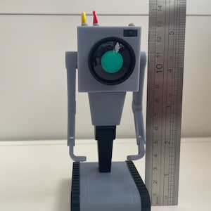 Puede incluir: Un juguete robot gris con cabeza cuadrada, brazos y base de orugas. Tiene una lente circular grande y dos antenas. El robot mide aproximadamente 25 cm de altura, medido con una regla.
