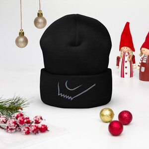 May include: A black beanie with a white embroidered design on the cuff. The beanie is displayed with holiday ornaments and decorative figurines. The beanie is a winter accessory.