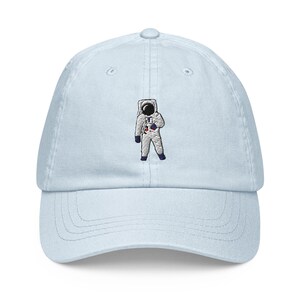 May include: A light blue baseball cap with an embroidered astronaut design. The astronaut is wearing a white spacesuit with a black helmet and a red patch on the chest.