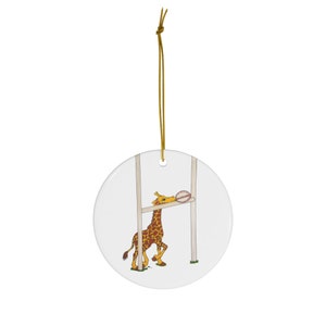 May include: A ceramic Christmas ornament featuring a cartoon giraffe scoring a try in a rugby match. The giraffe is brown and yellow with a pink rugby ball in its mouth. The ornament is white with a gold string.