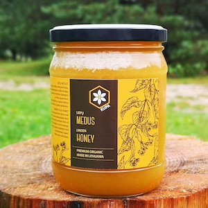 Peut inclure: Un pot de miel de tilleul avec un couvercle noir, posé sur une souche d'arbre. L'étiquette indique "LIEPŲ MEDUS LINDEN HONEY" avec un motif floral et "PREMIUM ORGANIC MADE IN LITHUANIA". Le miel est de couleur ambre doré.