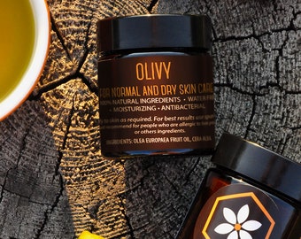 May include: Two small glass jars with brown lids. The jar on the left is labeled "OLIVY FOR NORMAL AND DRY SKIN CARE 100% NATURAL INGREDIENTS - WATER FREE - MOISTURIZING - ANTIBACTERIAL" and the jar on the right is labeled "KUPA". The jars are sitting on a wooden surface with green olives.