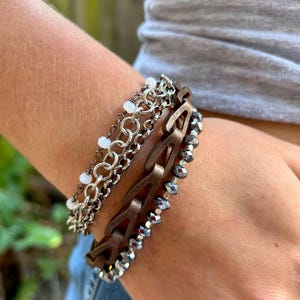 May include: A layered bracelet set featuring a brown leather bracelet with a chain link design, a silver chain bracelet with small white beads, and a black beaded bracelet.