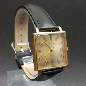 Puede incluir: Un reloj de pulsera cuadrado dorado con una correa de cuero negro. La esfera del reloj tiene marcadores de hora en números romanos y una aguja de segundos dorada.