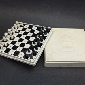 Könnte beinhalten: Ein weiß-schwarzes faltbares Schachspiel mit einem weißen Kunststoffkoffer. Der Koffer hat ein erhabenes Design eines Schiffes mit dem Text "La Navicella" darauf.