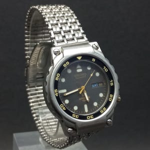 May include: A silver Seiko Quartz watch with a black dial and a stainless steel strap. The watch face shows the day and date, with luminous markers and hands. The bezel has black numerals and markings.