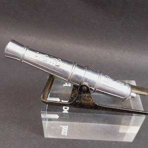 Könnte beinhalten: Ein silberfarbenes Metallkanonenmodell mit eingravierten dekorativen Schnörkeln entlang des Laufs. Die Kanone ist auf einem dunklen, verwitterten Metallgestell montiert, das eine Höhenverstellung ermöglicht und auf einem klaren Acrylblock ruht.