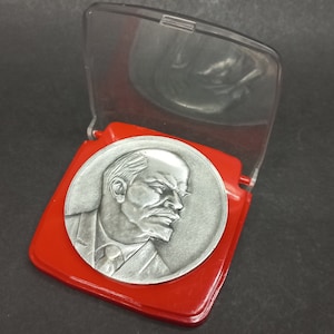 以下が含まれることがあります： スーツを着た男性の横顔が刻まれた銀貨。赤いプラスチック製のケースに透明なプラスチック製の蓋が付いています。