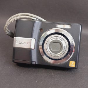 Puede incluir: Una cámara digital Lumix de color gris oscuro con una correa de muñeca plateada. La parte frontal presenta un objetivo Lumix DC Vario con zoom óptico de 3x, un flash y un pequeño logotipo cuadrado amarillo con una L blanca sobre una placa.