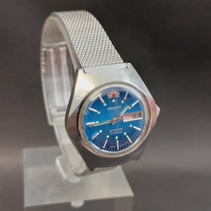 Op de afbeelding: Een zilverkleurig Orient automatisch polshorloge met een ronde blauwe gradiënt wijzerplaat, zilveren uurmarkeringen en een dag-datumvenster op de drie-uurpositie. Het horloge heeft een roestvrijstalen mesh-band en een gepolijste metalen kast.