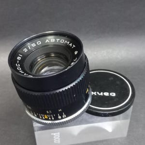 May include: Black camera lens with silver and yellow markings, and a black lens cap. The lens has the text "2/50 AUTOMAT" and "MOC-81" printed on it. The lens cap has the text "Kиев" printed on it.